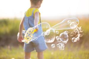 en flicka i en jeansoverall blåser såpbubblor på sommaren i ett fält vid solnedgången. internationella barndagen, glada barn, utomhusaktiviteter. sommar bakgrund. hälsosam och miljövänlig livsstil foto