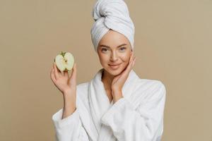 vacker frisk ung kvinna poserar med badlakan på huvudet klädd i morgonrock håller hälften av äpplet tittar direkt på kameran utergoes ansiktsbehandlingar efter att ha tagit dusch poser inomhus. foto