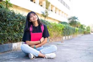 student ung flicka med rosa ryggsäck sitter vid foten med skolbakgrund. foto
