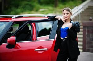 porträtt av vacker blond sexig modekvinna modell i mössa och i helsvart, blå korsett, med ljus smink nära röd stadsbil. foto