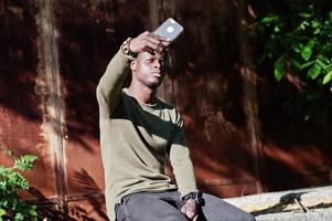 porträtt av en svart afrikansk amerikansk kille som sitter på det gamla staketet och använder sin smartphone. foto