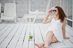 porträtt av en fantastisk ung kvinna i solglasögon och klänning som sitter på vitt trägolv med sin drink. foto