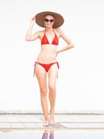 sexig tjej glad tjej i röd bikini bär hatt och solglasögon nära poolen med vit vägg. foto