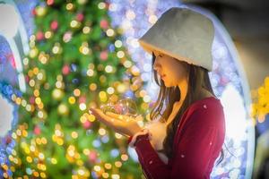 asiatisk vacker kvinna i röd vit skjorta som bär en vit hatt står lyckligt. i hennes hand höll han en lampa medan hon står framför julgranen med bokeh som bakgrund i temat foto