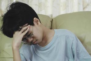 liten asiatisk pojke som bär glasögon med handen på huvudet, huvudvärk på grund av stress. han ledsen med lidande migrän. foto