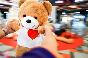 ljusbrun björndocka som bär vit t-shirt med rött hjärta på, svängs av mänsklig hand foto