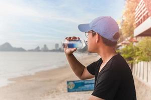 man som dricker vatten från en flaska sitta utanför bredvid stranden. foto