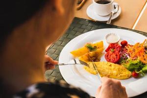 kvinna hand som håller gaffel och kniv äter frukost med omelett potatis, tomater och grönsakssallad på bordet. foto