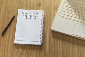 text på anteckningsblock med bärbar dator och penna på träskrivbord - checklista för ansökan om handelslicens foto