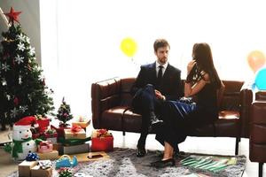 söta par älskar leende och tillbringar romantisk jultid och firar nyårsafton på brun soffdekoration med julgran, färgglad ballong och presentförpackningar i vardagsrummet hemma foto