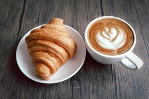 nybakad läcker croissant och en kopp vackert morgonkaffe på frukostbordet i trä. foto