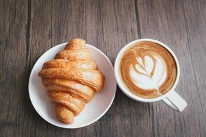 nybakad läcker croissant och en kopp vackert morgonkaffe på frukostbordet i trä, ovanifrån foto