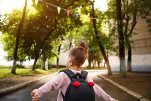 en liten flicka av kaukasiska i en skoluniform med en ryggsäck tittar på vägen på skolgården. koncept tillbaka till skolan. grundskola, utveckla verksamhet för förskolebarn. utrymme för text foto