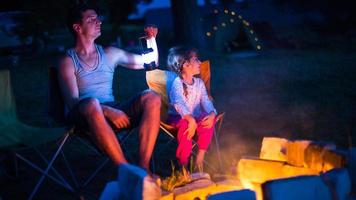 pappa och dotter sitter på natten vid brasan i det fria på sommaren i naturen. familjecamping, samlingar kring lägerelden. fars dag, grillning. campinglykta och tält foto