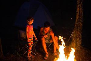 pappa och dotter sitter på natten vid brasan i det fria på sommaren i naturen. familjecamping, samlingar kring lägerelden. fars dag, grillning. campinglykta och tält foto