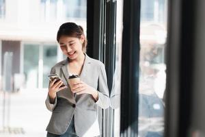 asiatisk kvinna med smartphone stående mot gata suddig byggnad bakgrund, mode affärsfoto av vacker flicka i avslappnad svit med smart telefon. foto