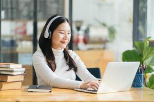 leende kvinna som bär trådlösa hörlurar arbetar med att skriva på anteckningsboken sitta vid skrivbordet på kontorsarbetsplatsen. njut av e-lärandeprocess, enkel bekväm applikationsanvändning, lyssna på musik under arbetsdagens koncept foto