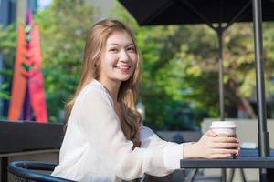 ung asiatisk vacker kvinna som bär kostym med bronshår sitter leende på stolen i kaféet utomhus medan hon håller kaffekoppen i handen på en solig morgon. foto