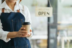 porträtt av glad kvinna som står vid dörröppningen till hennes butik. glad mogen servitris som väntar på kunder på kaféet. framgångsrik småföretagare i casual klädd i blått förkläde stående vid ingången foto