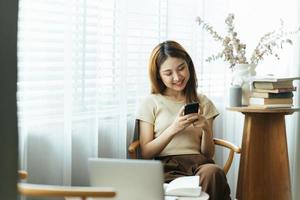 asiatisk kvinna i vardagskläder är glad och glad medan hon kommunicerar med sin smartphone och arbetar på ett kafé. foto