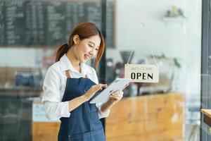 porträtt av glad kvinna som står vid dörröppningen till hennes butik. glad mogen servitris som väntar på kunder på kaféet. framgångsrik småföretagare i casual klädd i blått förkläde stående vid ingången foto