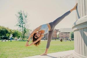 flexibel smal kvinna gör stretchövningar utomhusvistelse i god fysisk form klädd i cropped top och leggings står på ett ben lutar sig med armarna upphöjda värmer upp innan konditionsträning foto