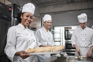 porträtt av afrikansk amerikansk kvinnlig kock i vit matlagningsuniform tittar på kameran med ett glatt leende och stolt med en bricka med bröd i köket, professionella bakverk och färskt bageriyrke. foto