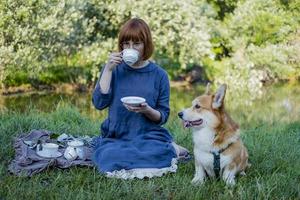 ung kvinna i retroklänning med rolig corgihund på picknicken, hona med söt hund dricker engelskt te i parken foto