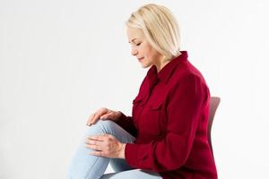 ledvärk, blond kvinna som rör vid benskada isolerad på vit bakgrund, kvinnlig reumatism, kvinna sitter på stol och rör vid knäsmärta foto
