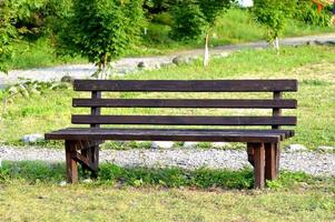 brun träbänk i parken. solig sommardag. grönt gräs och träd. vilo- och avkopplingsområde. tom bänk för att sitta. trä exteriört material. foto