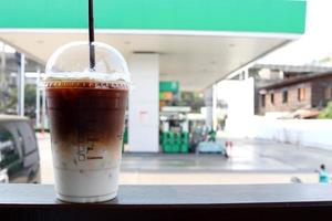 iskaffe i plastglas och halm på mörkbrunt bord och suddiga bensinstationbakgrund i thailand. foto