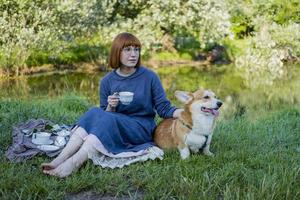 ung kvinna i retroklänning med rolig corgihund på picknicken, hona med söt hund dricker engelskt te i parken foto