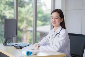 professionella asiatiska vackra unga leende kvinnliga läkare bär gummihandske sitter medan du väntar på patientundersökning på kontoret på sjukhuset. på bordet har papper, temperaturmätare och dator. foto