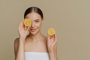 citron för skönhet och hälsa. vacker frisk ung kvinna täcker ögat med hälften av citronen njuter av spabehandling insvept i badhandduk isolerad över beige bakgrund kopieringsutrymme för din marknadsföring foto