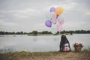 liten söt flicka sitter på långt grönt gräs utanför. flicka håller färgglada ballonger i handen. foto