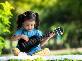 asiatisk liten flicka som sitter på mattan, koppla av och spelar ukulele utanför skolan för att njuta i naturparken foto