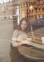 en vacker flicka sitter på café och tittar eftertänksamt ut genom fönstret. reflektion av staden i fönstret. brunett kvinna med långt hår ler och dricker cappuccino, vertikal foto
