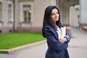 leende flicka brunett student håller anteckningsböcker och läroböcker, står vid universitetet på gatan i st. petersburg. en charmig kvinna med långt mörkt hår studerar på kurs och skaffar sig utbildning foto