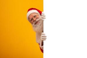 glad jultomte tittar ut från bakom den tomma skylten isolerad på gul bakgrund med kopia utrymme foto