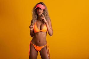 attraktiv lockigt hår flicka i en orange bikini, hatt, känslomässigt öppnad mun på en gul bakgrund med en perfekt kropp. isolerat. studio skott. foto