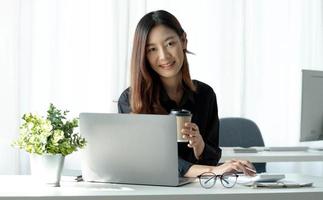 leende asiatisk affärskvinna som håller en kaffemugg och bärbar dator på kontoret. tittar på kameran. foto