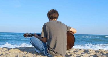 ung stilig man spelar i akustisk gitarr på stranden i solig dag, havet eller havet på bakgrunden foto