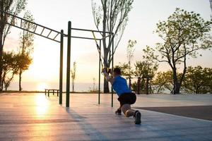 träning med upphängningsremmar i utegymmet, stark man som tränar tidigt på morgonen i parken, soluppgång eller solnedgång i havsbakgrunden foto