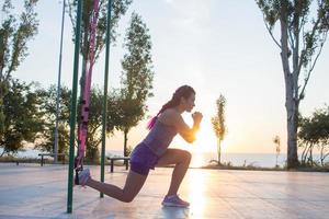 vacker passform kvinna i rosa och lila sportkläder träning på utomhusgym på morgonen, övningar med upphängningsremmar i parken foto