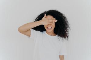 ung tonåring dam täcker ögonen med handflatan, gömmer sig, ler tandigt, har afro buskig frisyr, bär vit mockup t-shirt, står ensam i studion, väntar på överraskning, upprymd av förväntan foto