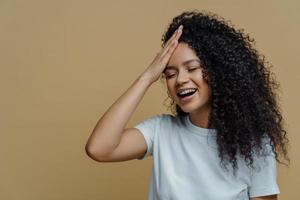 horisontell bild av glad positiv afroamerikansk kvinna har roligt, håller handen på pannan, fnissar glatt, sluter ögonen, har buskigt lockigt hår, bär casual vit t-shirt, isolerad på beige bakgrund. foto