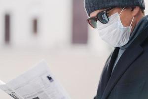 beskuren bild av manlig modell skyddar mot smittsam sjukdom eller coronavirus, bär hygienisk mask för att förhindra infektion, luftvägssjukdom, läser tidning, poserar utomhus i karantän stad foto