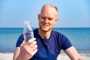 ung manlig idrottare dring färskvatten från plastflaska på stranden foto