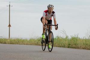 cykelracer i hjälm och sportkläder träning ensam på tom landsväg, fält och träd bakgrund foto