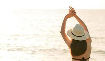 bakifrån av glad ung asiatisk kvinna i svart bikini och halmhatt slappna av och njut av den tropiska paradisstranden i solnedgången. flicka i sommarlovet mode. sexig modell. elegant livsstil. sommarvibbar. foto